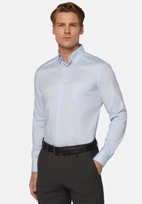 Svetlomodrá košeľa s dlhými rukávmi, vreckom na hrudi a golierom, vyrobená z hladkej látky. Skombinovaná s tmavosivými nohavicami.