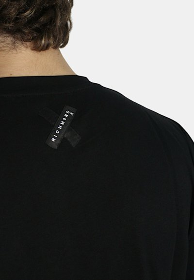 T-shirt nera realizzata in tessuto morbido con colletto rotondo. Presenta un'etichetta nera rettangolare sul retro con testo bianco e un design a croce.