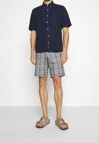 Korte mouwen marineblauw button-up shirt met zak, gecombineerd met grijs geruite shorts en bruine sandalen met dubbele banden en een gestructureerde afwerking.