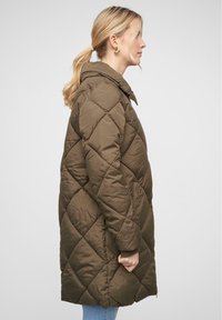 Manteau long vert olive matelassé avec capuche, présentant un piquage en forme de diamant, des poches latérales et des poignets côtelés. Texture douce et design décontracté.