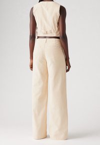 Nainen, joka on pukeutunut beigeihin leveälahkeisiin housuihin ja ilman hihoja olevaan lyhytlihaiseen liiviin, nähtynä takaa, yksinkertaista vaaleaa taustaa vasten.