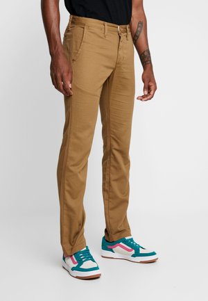 Mann trägt beige gerade geschnittene Hose und weiße Sneakers mit türkis- und pinkfarbenen Akzenten, steht vor einem einfarbigen grauen Hintergrund.