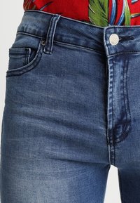 Blaue Jeans aus Denim mit mittelhohem Bund, Vordertaschen und einer dezenten, ausgewaschen wirkenden Textur sowie einem silbernen Knopfverschluss.