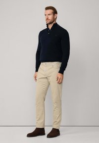 Pull vert foncé avec un col zippé, associé à un pantalon chino beige et des bottes chukka marron. Texture lisse, coupe ajustée et détails minimalistes.
