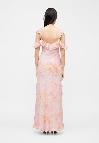 Γυναίκα με μακριά σκούρα μαλλιά, ντυμένη με μακρύ ροζ φόρεμα με λουλουδάτο σχέδιο και βολάν στους ώμους, με την πλάτη προς τον θεατή.