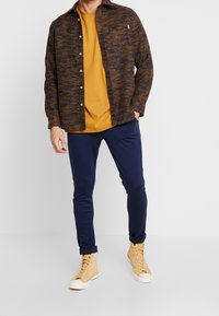 Hnědá vzorovaná košile přes hořčicové tričko, kombinovaná s námořnickými kalhotami a béžovými vysokými teniskami. Knoflíková košile má kapsu.