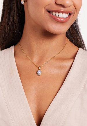 Gouden ketting met een ronde, lichtblauwe stenen hanger omringd door kleine witte stenen, tentoongesteld op een textuur, lichtkleurige stof.