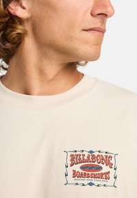 Light beige short-sleeve shirt featuring a colorful graphic logo on the chest. The logo includes red and blue text with decorative borders.