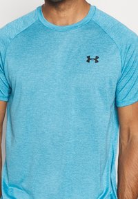 Man med kortärmad, ljusblå, atletisk T-shirt med en svart Under Armour-logotyp på vänster bröst.