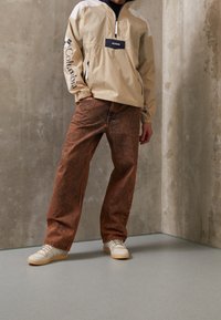 Beige uppdragströja med marinblå detaljer och dragkedja, i kombination med bruna strukturerade jeans och vita sneakers. Enkelt design med en avslappnad passform.