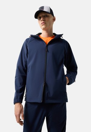 Młody mężczyzna w granatowej kurtce na zamek, pomarańczowej koszulce, granatowych spodniach i białej oraz granatowej czapce z daszkiem, stojący na tle białego tła.