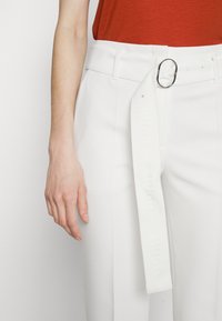 Witte hoge taillebroek met een brede riem met een ronde gesp en reliëf logo-detail, aangevuld met een gestructureerde rode top.