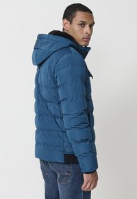 Chaqueta acolchada azul con capucha, con textura de acolchado, puños elásticos y bolsillos frontales. Logo de la marca embosssed en la parte posterior.