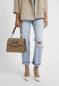 Ruskeakuvioinen käsilaukku, jossa on jämäkkä muoto, kullanväriset metalliosat ja lyhyt kahva. Yhdistetty vaaleansinisiin repeytyneisiin farkkuihin ja valkoisiin korkokenkiin.