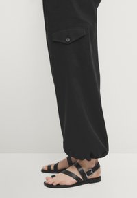 Mustat löysät housut, joissa on sivutasku ja napin yksityiskohta, yhdistetty mustiin tasapohjaisiin sandaaleihin, joissa on useita hihnoja ja hopeinen solki.