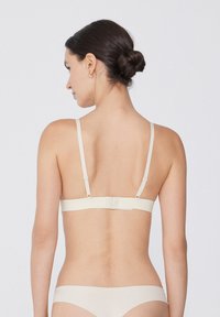 Beige bralette s nastavitelnými popruhy a háčkovým zapínáním, se hladkým povrchem a minimalistickým designem. Pohled zezadu zdůrazňuje jednoduchost.