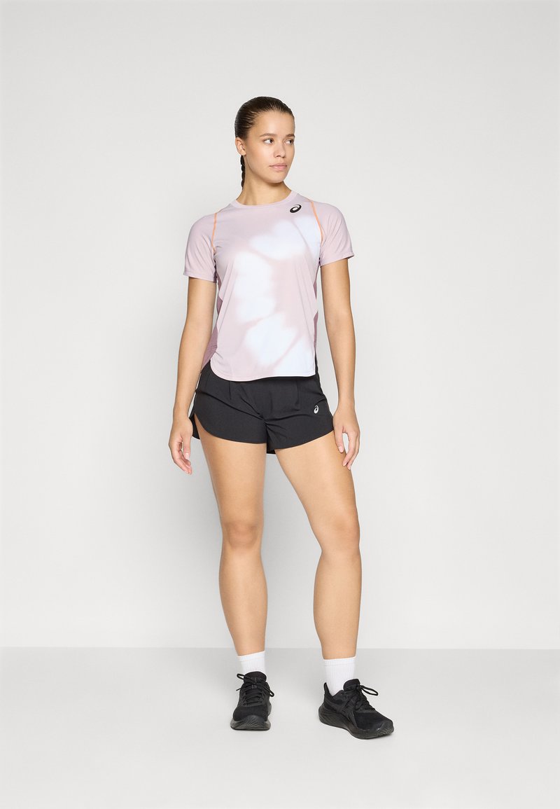 Helles rosa Sportoberteil mit kurzen Ärmeln und orangefarbenen Akzenten, kombiniert mit schwarzen Sportshorts. Das Model trägt schwarze Sneakers und weiße Socken.