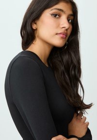 Junge Frau mit langen, dunklen Haaren, die große goldene Creolen und ein schwarzes Langarmoberteil trägt, und leicht seitlich schaut mit verschränkten Armen.