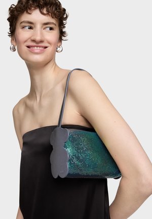 Joven sonriendo, vistiendo un top negro sin tirantes y llevando un bolso brillante color teal con un detalle lateral de cuero en forma de oso sobre el hombro.