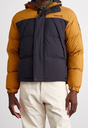 Homme portant une doudoune Timberland jaune moutarde et noire avec une fermeture éclair frontale et des boutons, associée à un pantalon écru.