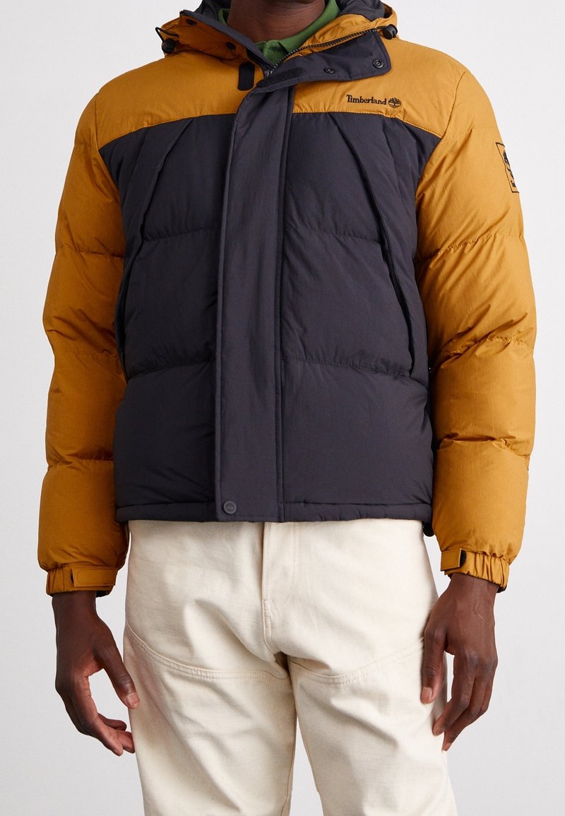 Homme portant une doudoune Timberland jaune moutarde et noire avec une fermeture éclair frontale et des boutons, associée à un pantalon écru.