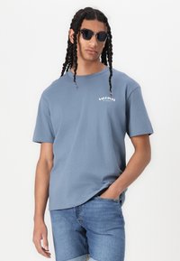 Lyseblå bomullst-skjorte med kort erm, med et hvitt "SARDINAS"-logo. Kombinert med denimshorts. Tilbehør inkluderer svarte solbriller.