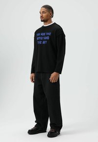 Schwarzes Langarmshirt mit blauem Text "DU BIST DER KÜNSTLER UND DAS KUNSTWERK", kombiniert mit schwarzen weiten Jeans und schwarzen Schuhen mit pinken Akzenten.