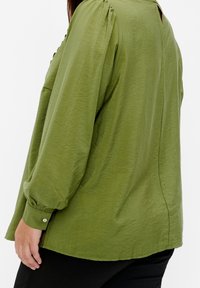 Kvinna i en lös, olivgrön blus med lång ärm och knäppta manschetter, tillsammans med svarta byxor, framför en vit bakgrund.