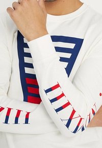 Camiseta blanca de manga larga con un diseño geométrico azul y rojo en la parte frontal y detalles en rojo en las mangas con numeración.