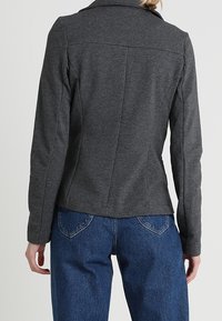 Tmavě šedý vypasovaný blazer s límečkem, přizpůsobenými švy a dlouhými rukávy. Kombinováno s modrými džínami s vysokým pasem, pohled zezadu.