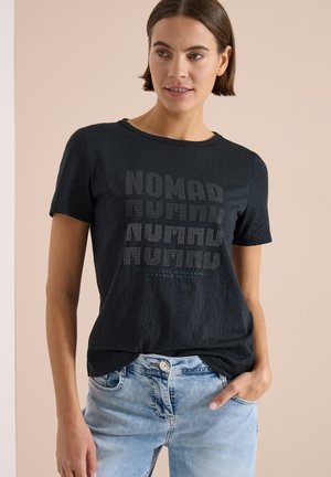 Jeune femme aux cheveux bruns courts portant un t-shirt noir à motif "NOMAD" et un jean bleu clair, debout devant un fond beige.