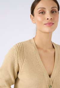 Beige gestrickte Strickjacke mit V-Ausschnitt, strukturiertem Schulterdetail und einer einzelnen Knopfleiste. Weicher Stoff mit glatter Oberfläche.