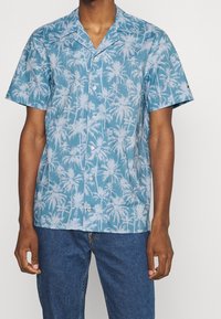 Camisa de manga corta en algodón azul claro con patrón de palmeras blancas, con cuello con solapa y cierre frontal con botones.