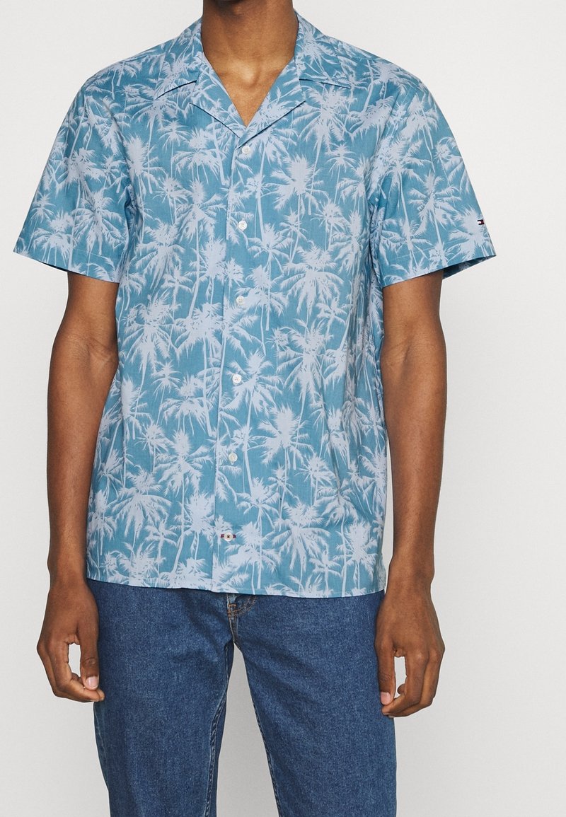 Camisa de manga corta en algodón azul claro con patrón de palmeras blancas, con cuello con solapa y cierre frontal con botones.