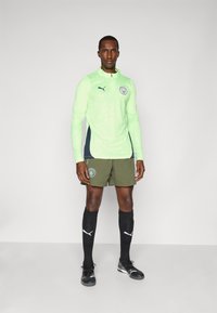 Lime žalios ilgos rankovės sportinis marškinėliai su tamsiai mėlynu šonu, alyvuogių spalvos šortai, juodos iki kelių kojinės ir juodi futbolo bateliai, ant marškinėlių yra logotipas.