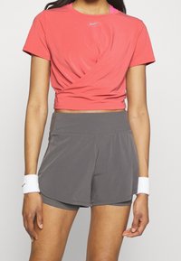 Top corto rojo con detalle torsionado en la parte delantera, combinado con pantalones cortos deportivos grises. Ambos hechos de un tejido ligero y transpirable. Incluye muñequeras blancas.