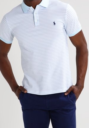 Mann trägt ein hellblau-weiß gestreiftes Poloshirt mit kleinem dunklem Logo auf der Brust, kombiniert mit dunkelblauer Hose, Hände in den Taschen.