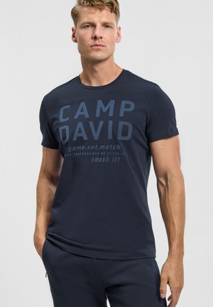 Blond man wearing a navy blue short-sleeve shirt with "CAMP DAVID" and other text, posing with one hand in his pocket on white background.