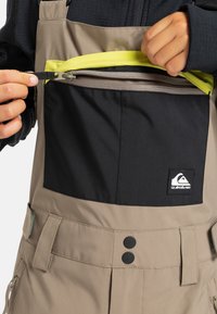 Salopette beige avec une poche zippée noire et une doublure jaune néon. Comprend deux boutons noirs et une étiquette de marque sur la poche.