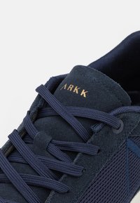 Námořnické tenisky vyrobené z nubuku a síťoviny, s plochými tkaničkami, vyšitým logem a texturovanými panely. Design zahrnuje diagonální linie a akcenty.