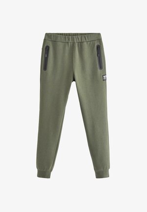 Lächelnder Junge steht nach vorne gerichtet, trägt ein schlichtes marineblaues T-Shirt, olivgrüne Jogginghosen und marineblaue Sneakers, die Hände in den Taschen.