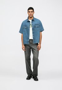 Lys blå denim kortermet skjorte med to brystlommer, brukt over en hvit t-skjorte med grafikk, kombinert med grå vide bukser og svarte sko.