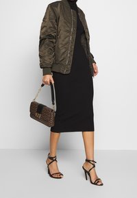 Kvinna som bär en olivgrön bomberjacka, svart midiklänning, svarta sandaletter med hög klack, håller en brun mönstrad handväska med guldkedja.