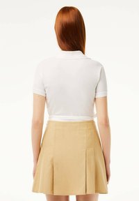 Maglietta polo bianca a maniche corte, abbinata a una gonna plisse beige, vista da dietro. Tessuto morbido, colletto classico e vestibilità sartoriale.