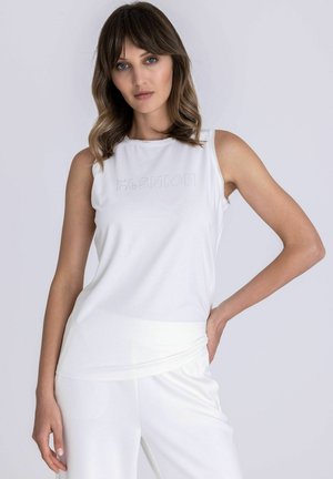 Femme aux cheveux bruns mi-longs, portant un haut blanc sans manches et un pantalon blanc, posant avec une main sur la hanche.