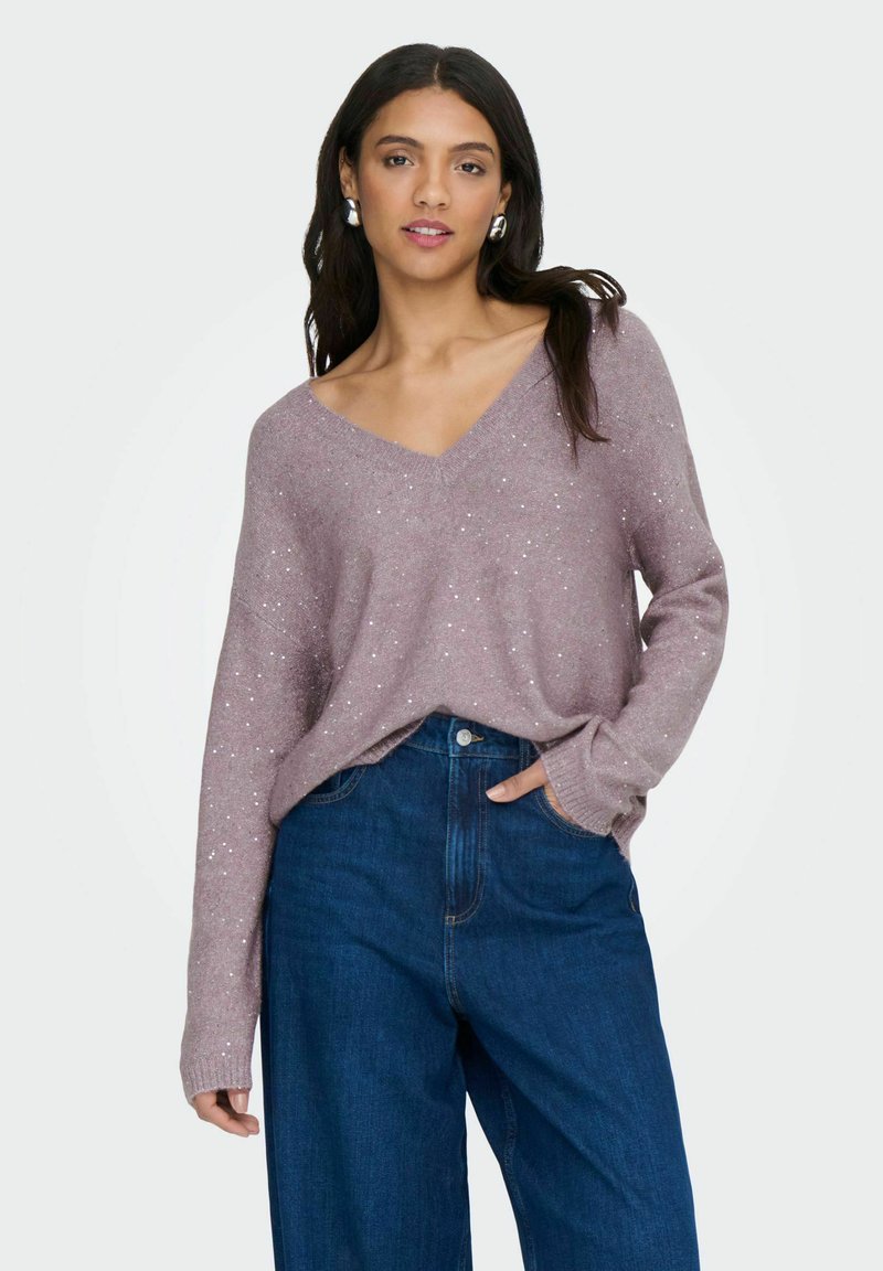 Mujer con un suéter lila claro de cuello en V con sutiles destellos y jeans oscuros de tiro alto, de pie con una mano en el bolsillo.