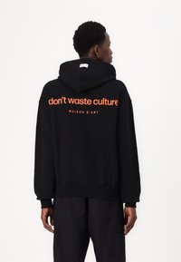Schwarzer Kapuzenpullover mit Kordelzug und Rippbündchen. Auf der Rückseite befindet sich orangefarbener Text: "verschwendet keine Kultur" und "MAISON D'ART."