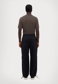 Man standing facing away wearing a brown short-sleeve shirt, black pants with back pockets, and white sneakers on a plain background.