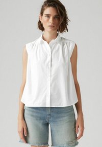 Vit ärmlös skjorta med knäppning framtill och samlad axeldetalj, tillsammans med ljusblå denimshorts med fransade kanter.