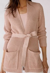 Cárdigan de punto rosa claro con un patrón texturizado, abertura frontal, cinturón anudado en la cintura y dos bolsillos delanteros. Mangas largas.